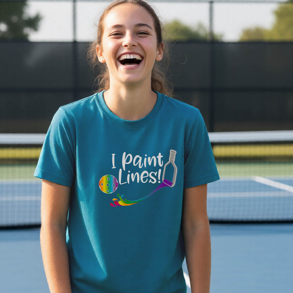 A young adult on an outdoor pickleball court wearing a teal short-sleeve t-shirt with a graphic that reads 'THIS IS MY PICKLEBALL SHIRT' in white with a yellow pickleball graphic on the back.
