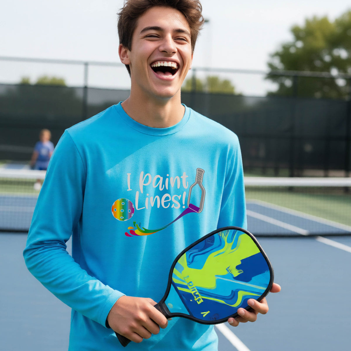 A close-up of a young adult wearing a bright blue long-sleeve t-shirt with a graphic on the front that has white text "I PAINT LINES'  with a white pickleball paddle painting multi-colored lines leading a mutli-colored pickleball.