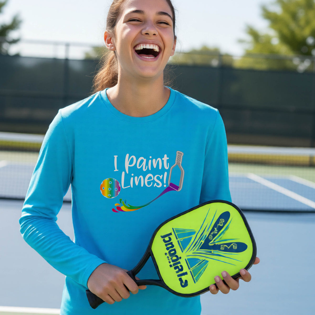 A close-up of a young adult wearing a bright blue long-sleeve t-shirt with a graphic on the front that has white text "I PAINT LINES'  with a white pickleball paddle painting multi-colored lines leading a mutli-colored pickleball.