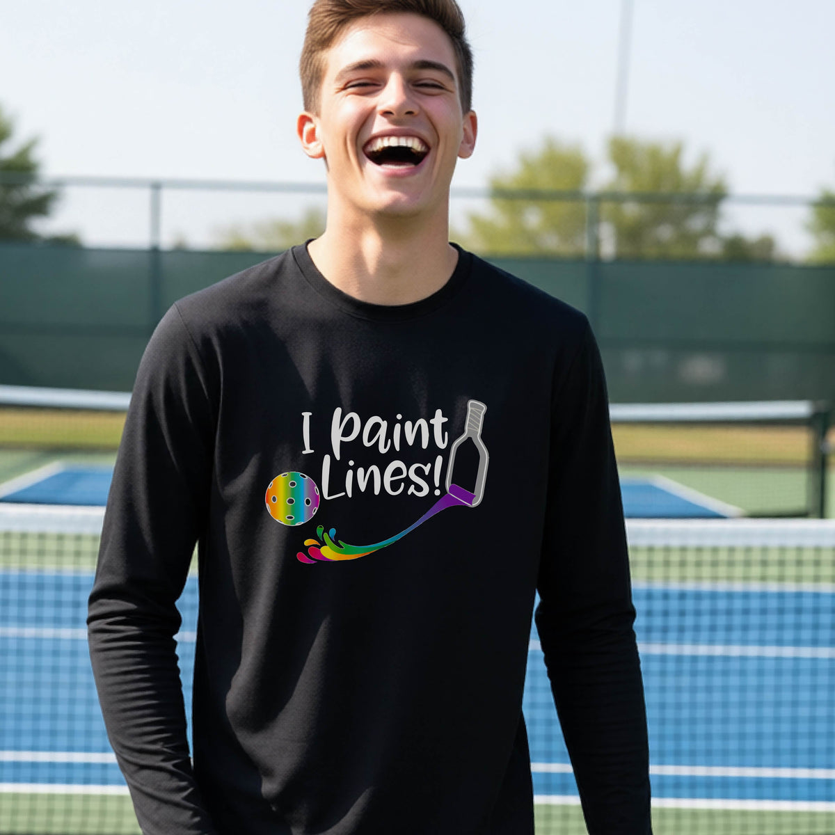 A close-up of a young adult wearing a black long-sleeve t-shirt with a graphic on the front that has white text "I PAINT LINES'  with a white pickleball paddle painting multi-colored lines leading a mutli-colored pickleball.