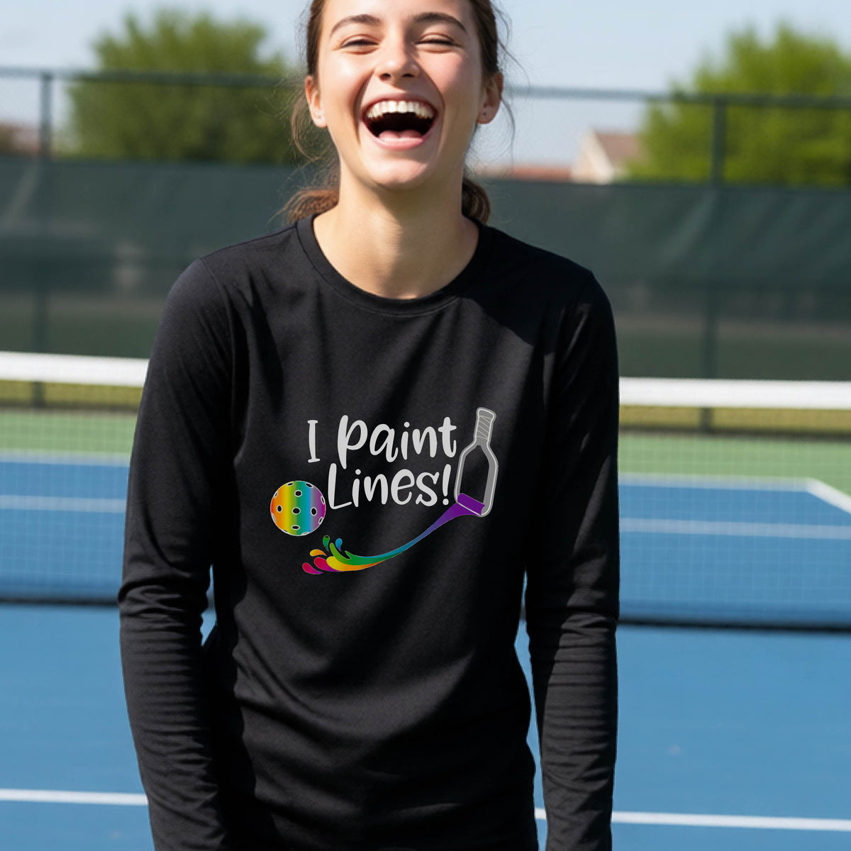 A close-up of a young adult wearing a black long-sleeve t-shirt with a graphic on the front that has white text "I PAINT LINES'  with a white pickleball paddle painting multi-colored lines leading a mutli-colored pickleball.