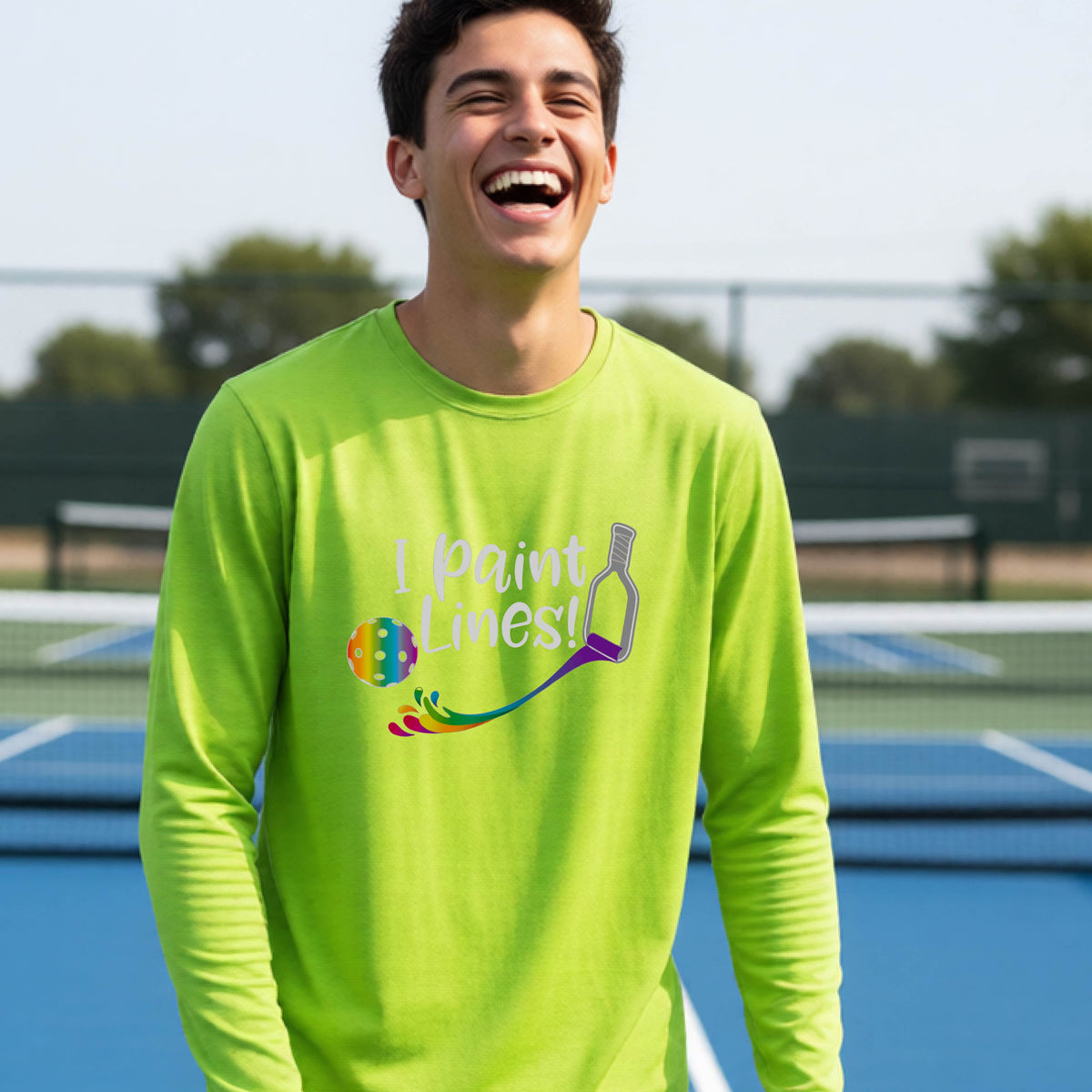 A close-up of a young adult wearing a lime green long-sleeve t-shirt with a graphic on the front that has white text "I PAINT LINES'  with a white pickleball paddle painting multi-colored lines leading a mutli-colored pickleball.