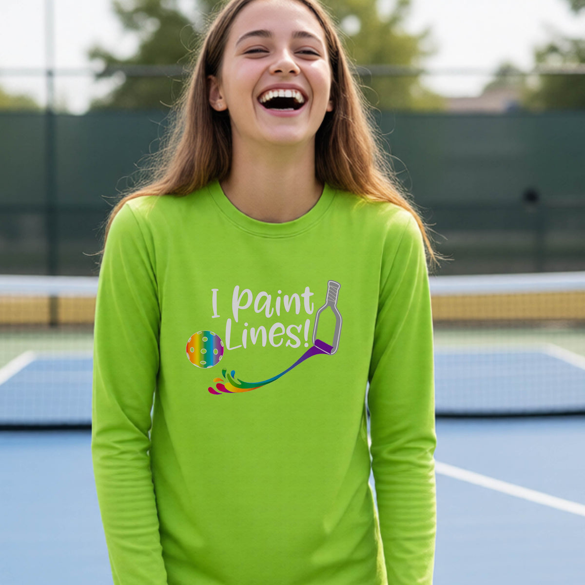 A close-up of a young adult wearing a lime green long-sleeve t-shirt with a graphic on the front that has white text "I PAINT LINES'  with a white pickleball paddle painting multi-colored lines leading a mutli-colored pickleball.