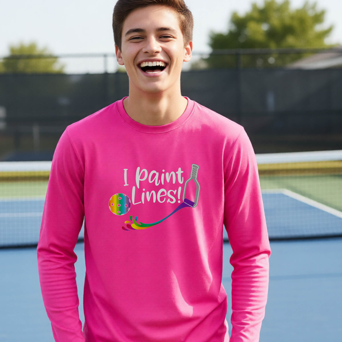 A close-up of a young adult wearing a pink long-sleeve t-shirt with a graphic on the front that has white text "I PAINT LINES'  with a white pickleball paddle painting multi-colored lines leading a mutli-colored pickleball.