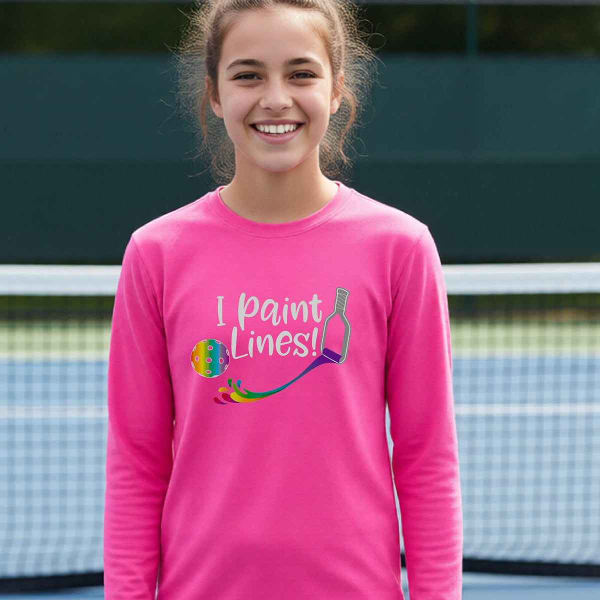 A close-up of a young adult wearing a pink long-sleeve t-shirt with a graphic on the front that has white text "I PAINT LINES'  with a white pickleball paddle painting multi-colored lines leading a mutli-colored pickleball.