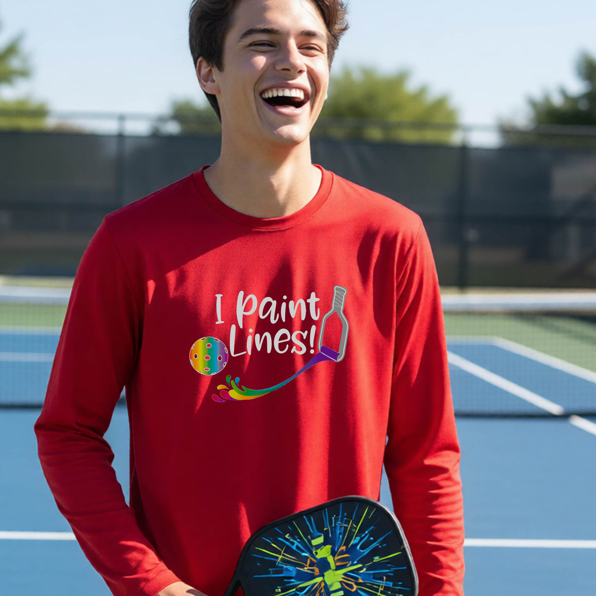 A close-up of a young adult wearing a red long-sleeve t-shirt with a graphic on the front that has white text "I PAINT LINES'  with a white pickleball paddle painting multi-colored lines leading a mutli-colored pickleball.