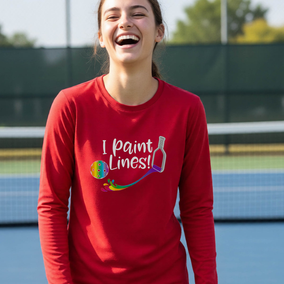 A close-up of a young adult wearing a red long-sleeve t-shirt with a graphic on the front that has white text "I PAINT LINES'  with a white pickleball paddle painting multi-colored lines leading a mutli-colored pickleball.