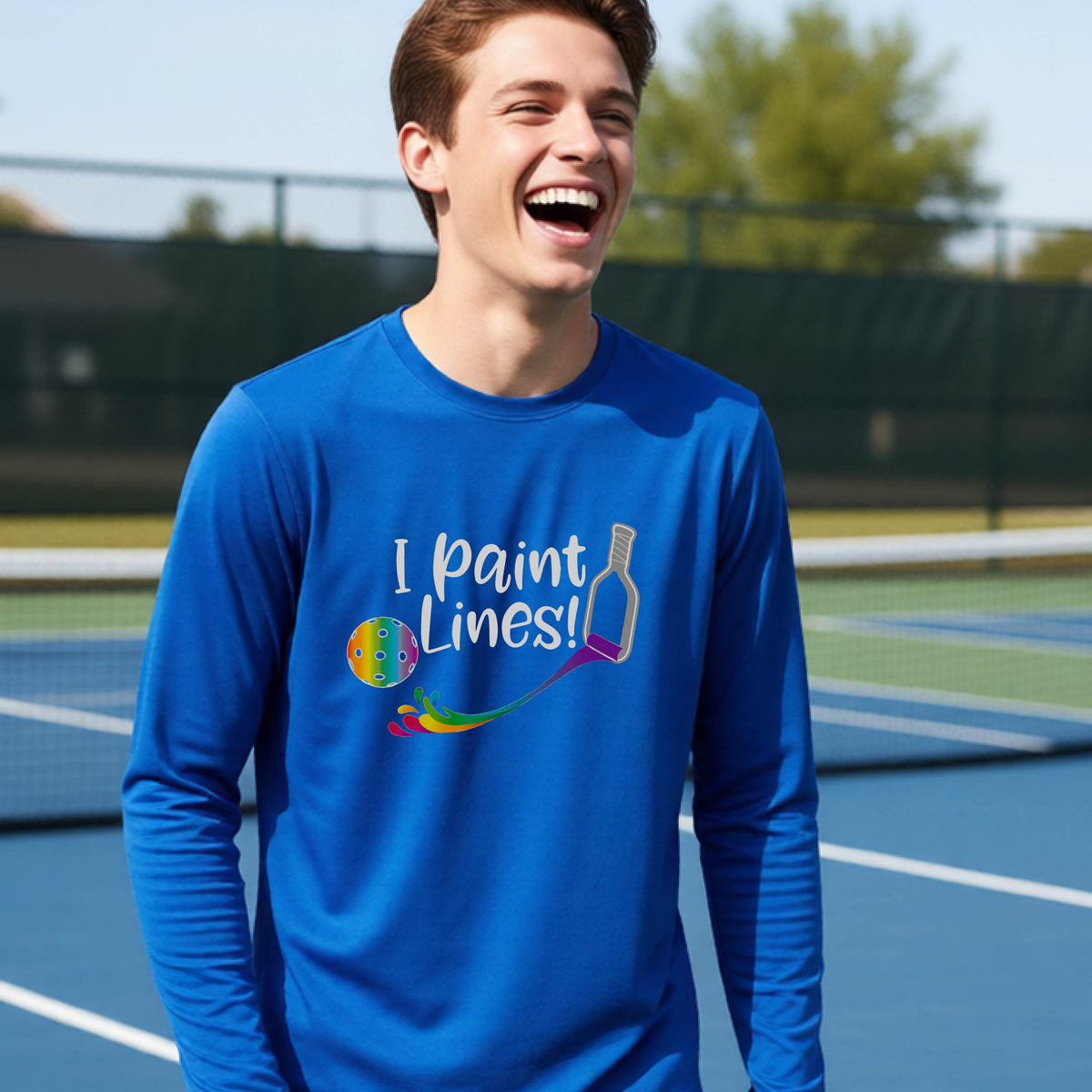 A close-up of a young adult wearing a royal blue long-sleeve t-shirt with a graphic on the front that has white text "I PAINT LINES'  with a white pickleball paddle painting multi-colored lines leading a mutli-colored pickleball.