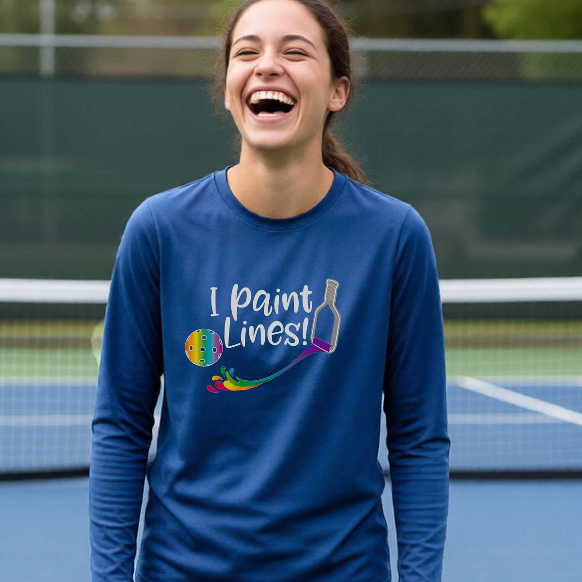 A close-up of a young adult wearing a royal blue long-sleeve t-shirt with a graphic on the front that has white text "I PAINT LINES'  with a white pickleball paddle painting multi-colored lines leading a mutli-colored pickleball.