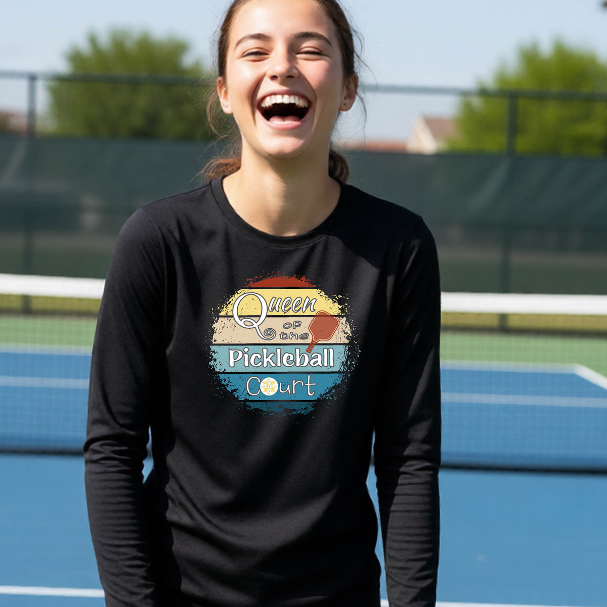 A woman laughing on an outdoor pickleball court wearing a black long-sleeve t-shirt with a retro-style graphic that reads 'Queen of the Pickleball Court' in a sunset gradient.
