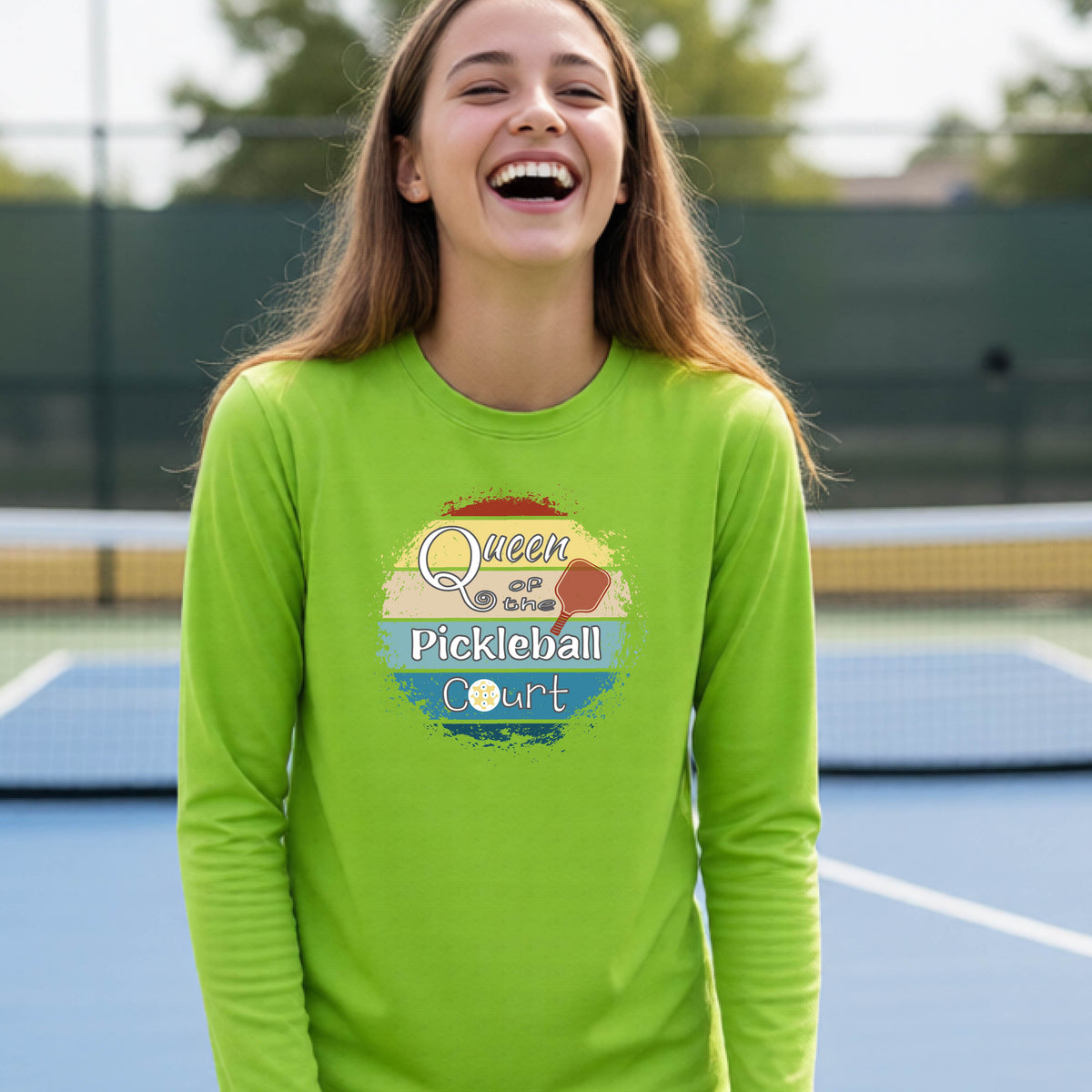A woman laughing on a blue pickleball court wearing a bright lime green long-sleeve t-shirt with a retro-style graphic that reads 'Queen of the Pickleball Court' in a retro sunset design.
