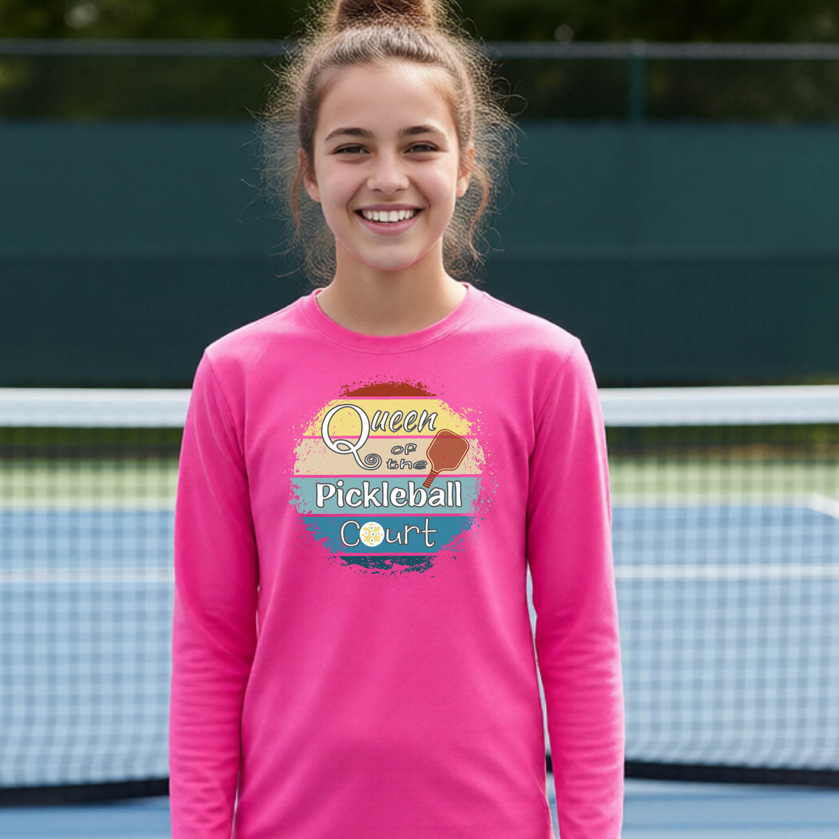 A smiling young woman stands on a pickleball court wearing a bright pink long-sleeve shirt with a colorful graphic that reads 'Queen of the Pickleball Court' over a retro sun and paddle design.