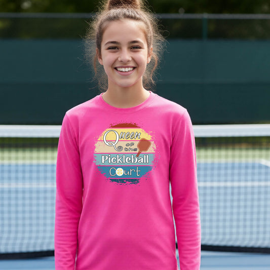 A smiling young woman stands on a pickleball court wearing a bright pink long-sleeve shirt with a colorful graphic that reads 'Queen of the Pickleball Court' over a retro sun and paddle design.