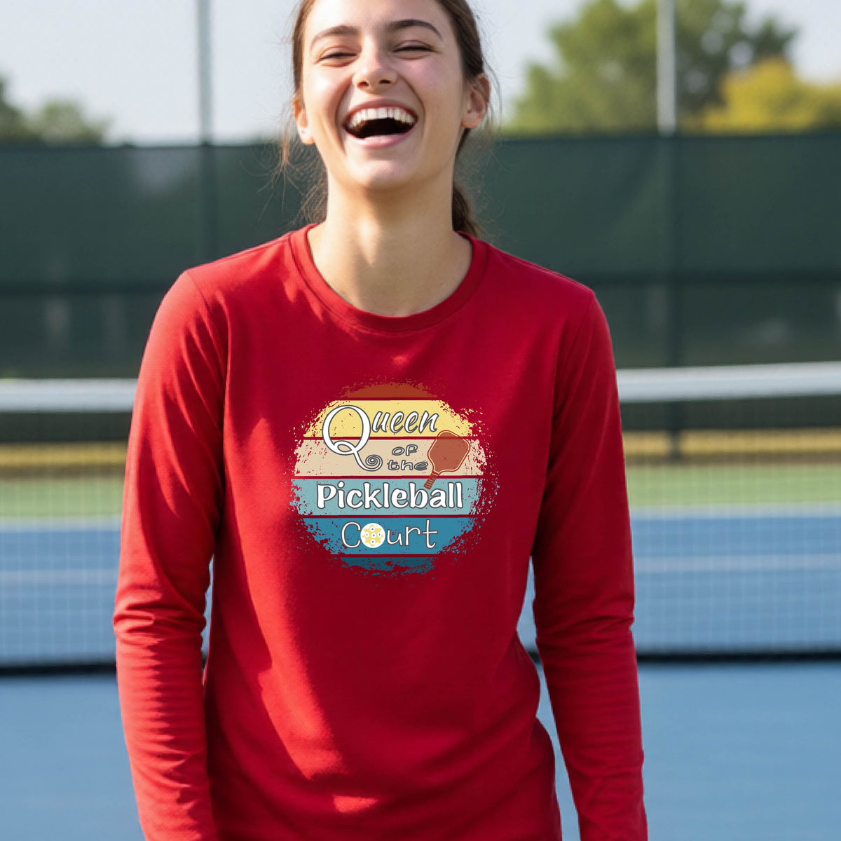 A woman laughing on a pickleball court wearing a red long-sleeve shirt with a retro-style graphic that reads 'Queen of the Pickleball Court' in yellow, beige, and blue.