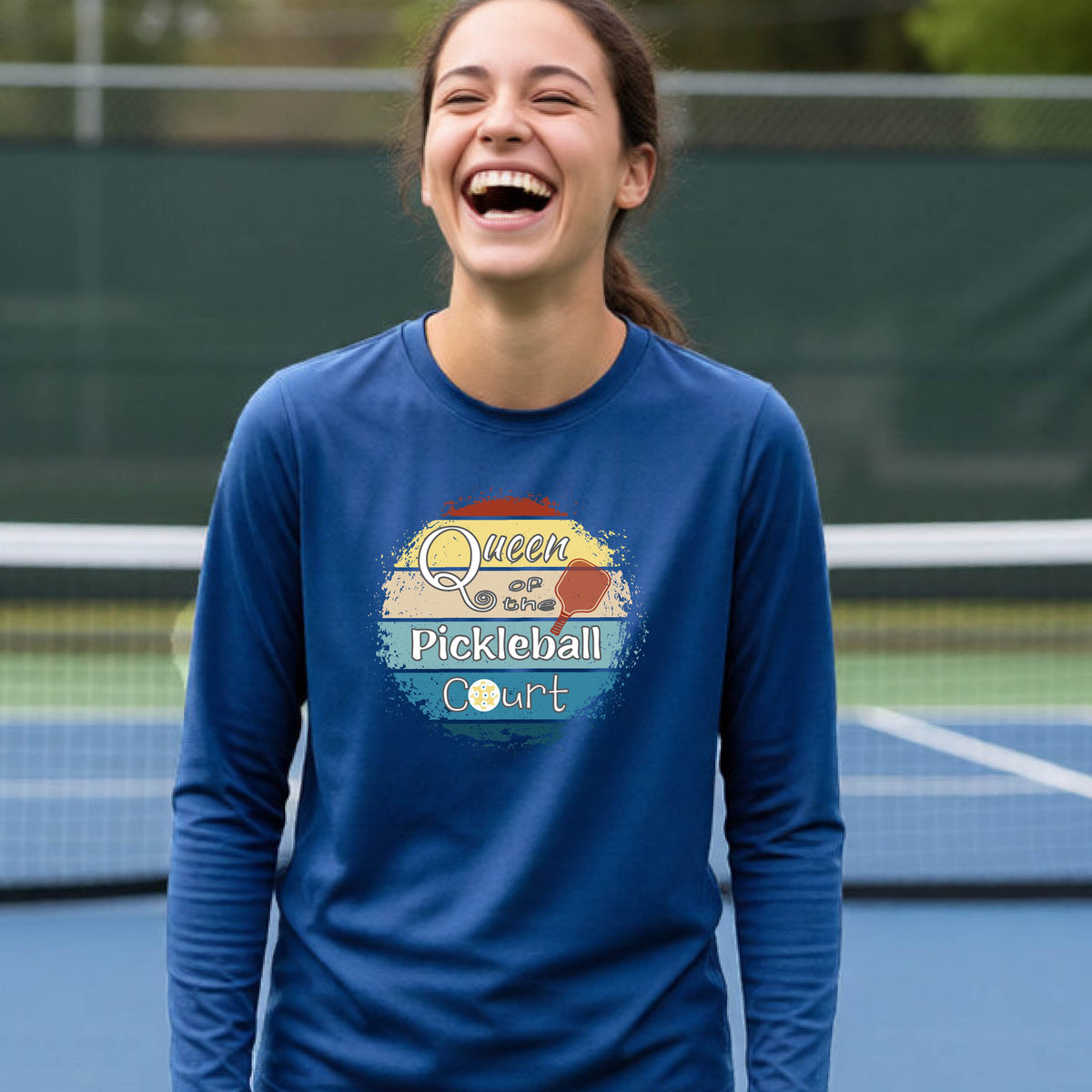 A woman laughing on a pickleball court wearing a royal blue long-sleeve t-shirt with a retro-style graphic that reads 'Queen of the Pickleball Court' over a vintage sunset  illustration.
