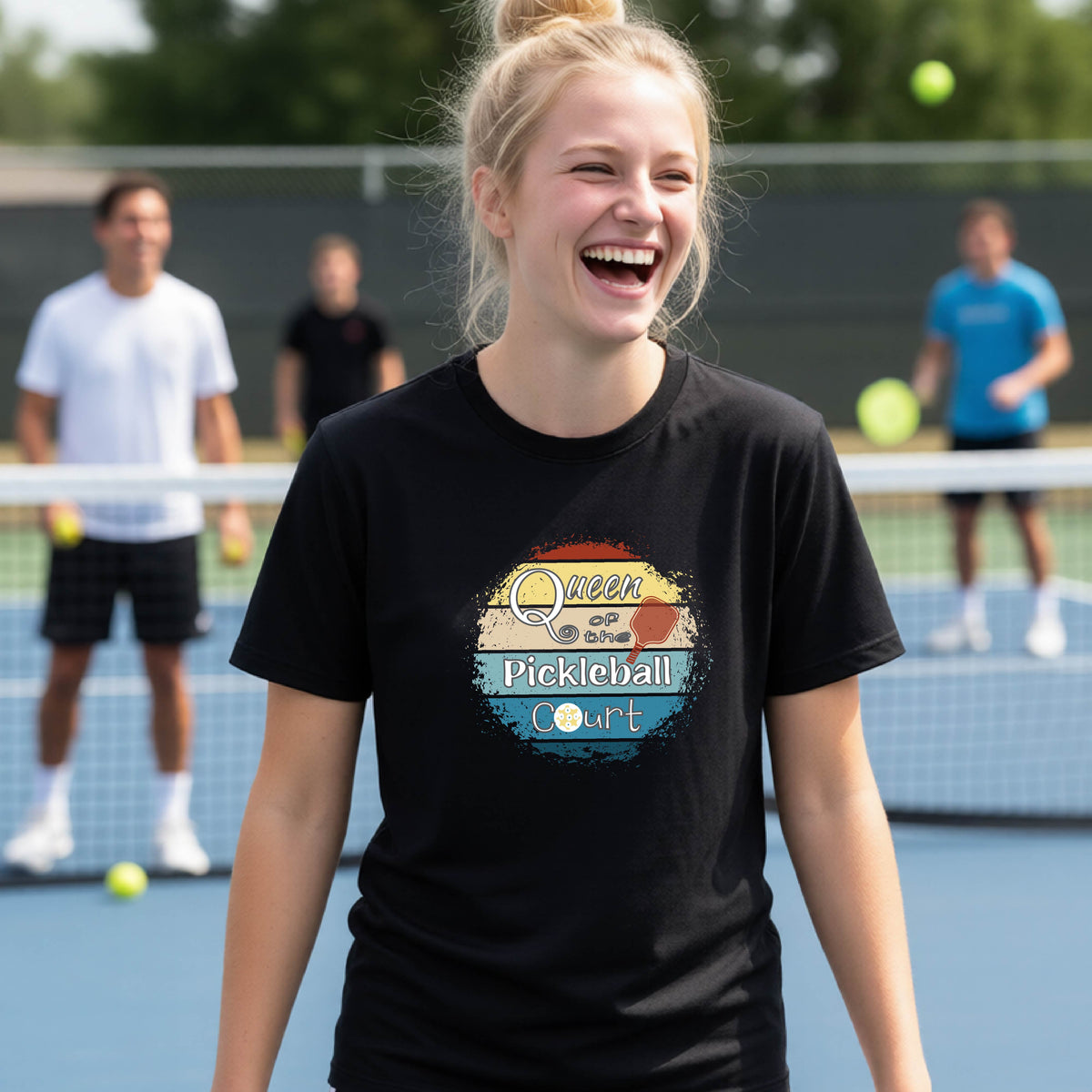 A young girl laughing on a pickleball court, wearing a black t-shirt with a retro-style graphic that reads 'Queen of hte Pickleball Court' in a yellow, orange, and blue retro sunset design.