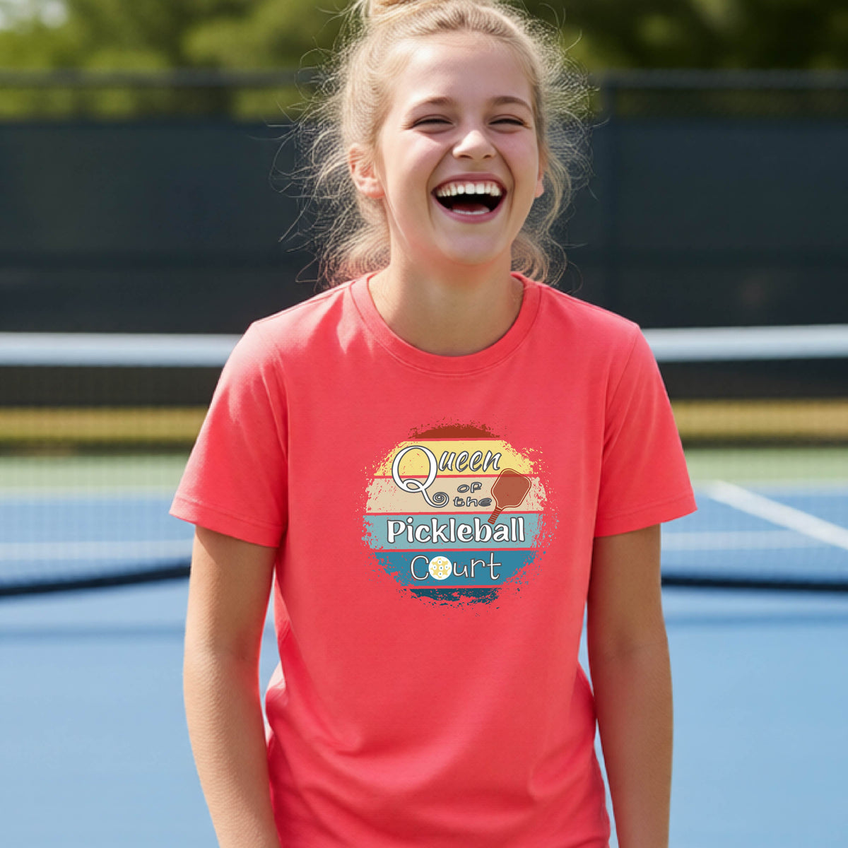 A young girl laughing on a pickleball court, wearing a coral pink t-shirt with a retro-style graphic that reads 'Queen of hte Pickleball Court' in a yellow, orange, and blue retro sunset design.