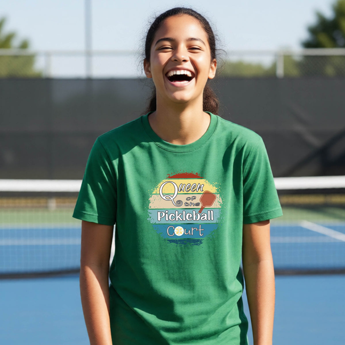 A young girl laughing on a pickleball court, wearing a green t-shirt with a retro-style graphic that reads 'Queen of hte Pickleball Court' in a yellow, orange, and blue retro sunset design.