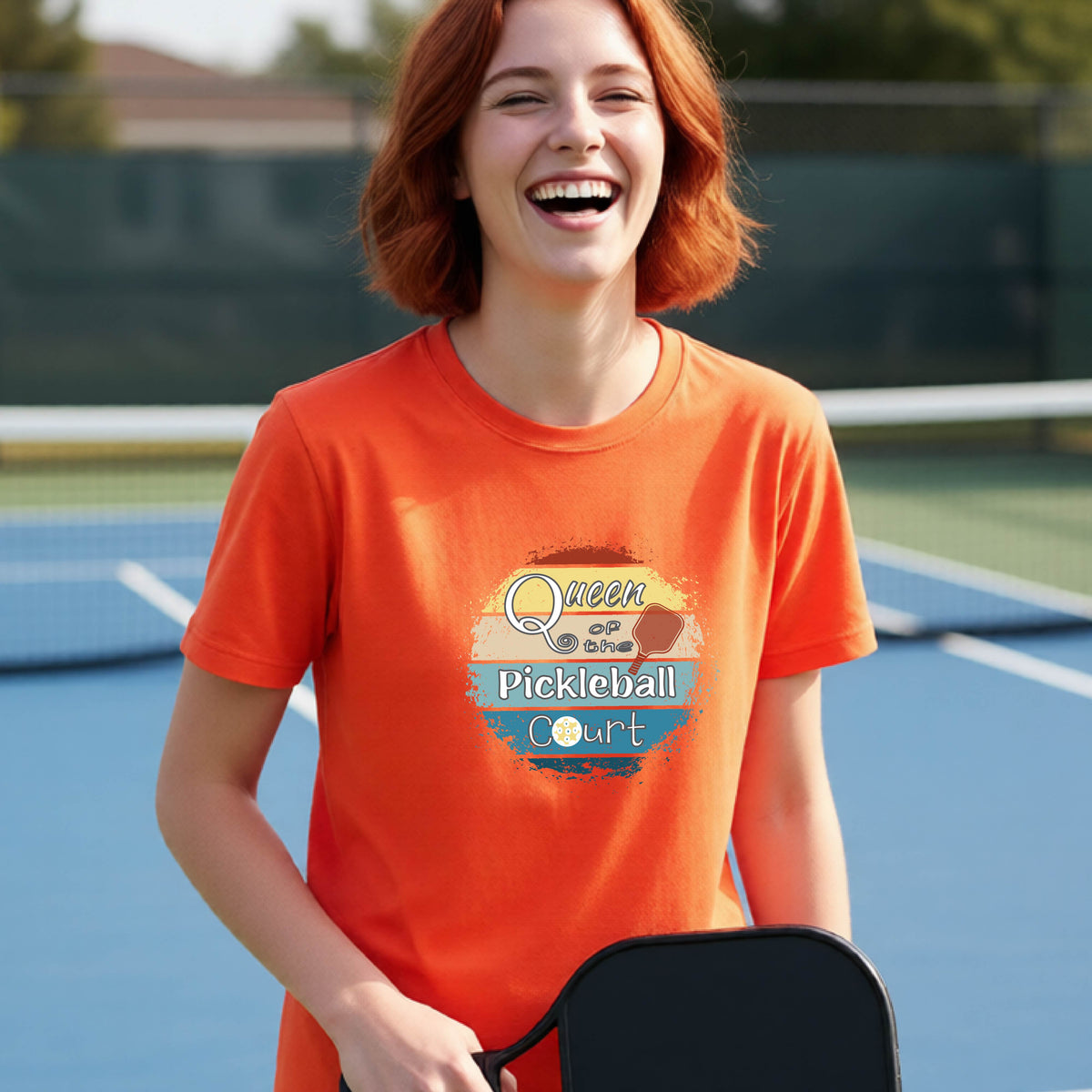 A young girl laughing on a pickleball court, wearing a bright orange t-shirt with a retro-style graphic that reads 'Queen of hte Pickleball Court' in a yellow, orange, and blue retro sunset design.