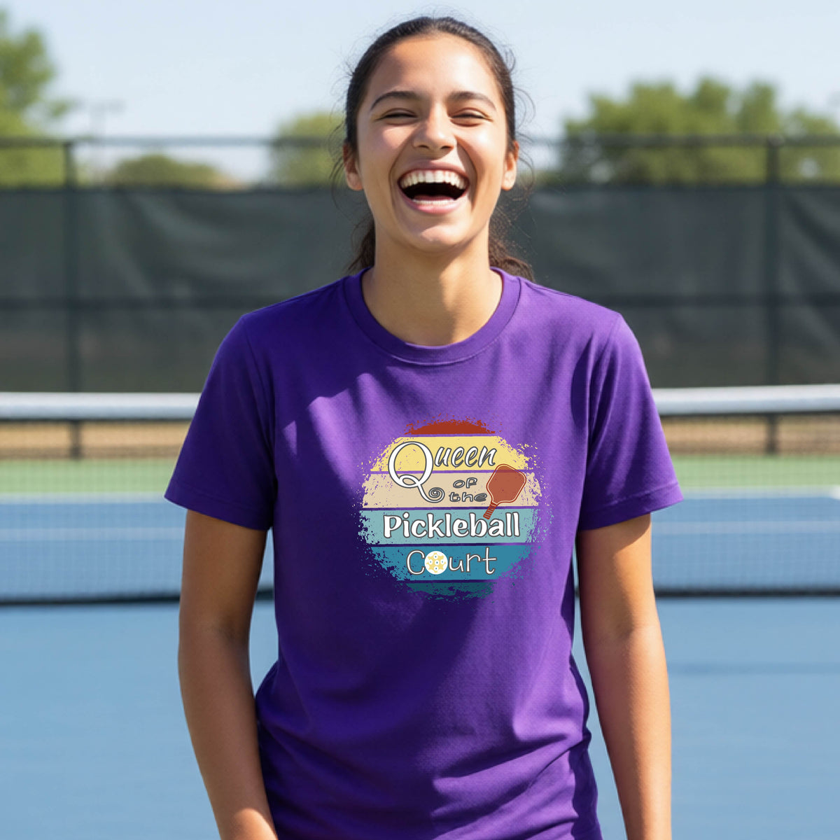 A young girl laughing on a pickleball court, wearing a purple t-shirt with a retro-style graphic that reads 'Queen of hte Pickleball Court' in a yellow, orange, and blue retro sunset design.