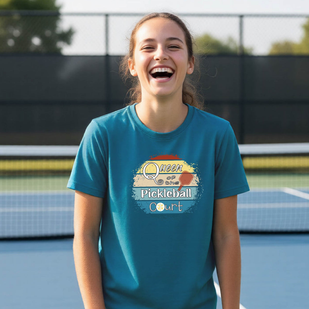 A young girl laughing on a pickleball court, wearing a teal t-shirt with a retro-style graphic that reads 'Queen of hte Pickleball Court' in a yellow, orange, and blue retro sunset design.