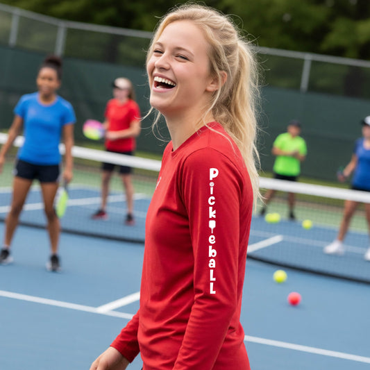 A young person wearing a red long-sleeve t-shirt with a white graphic text on the sleeve 'PICKLEBALL' with a pickleball paddle replacing the L, standing on a pickleball court. 