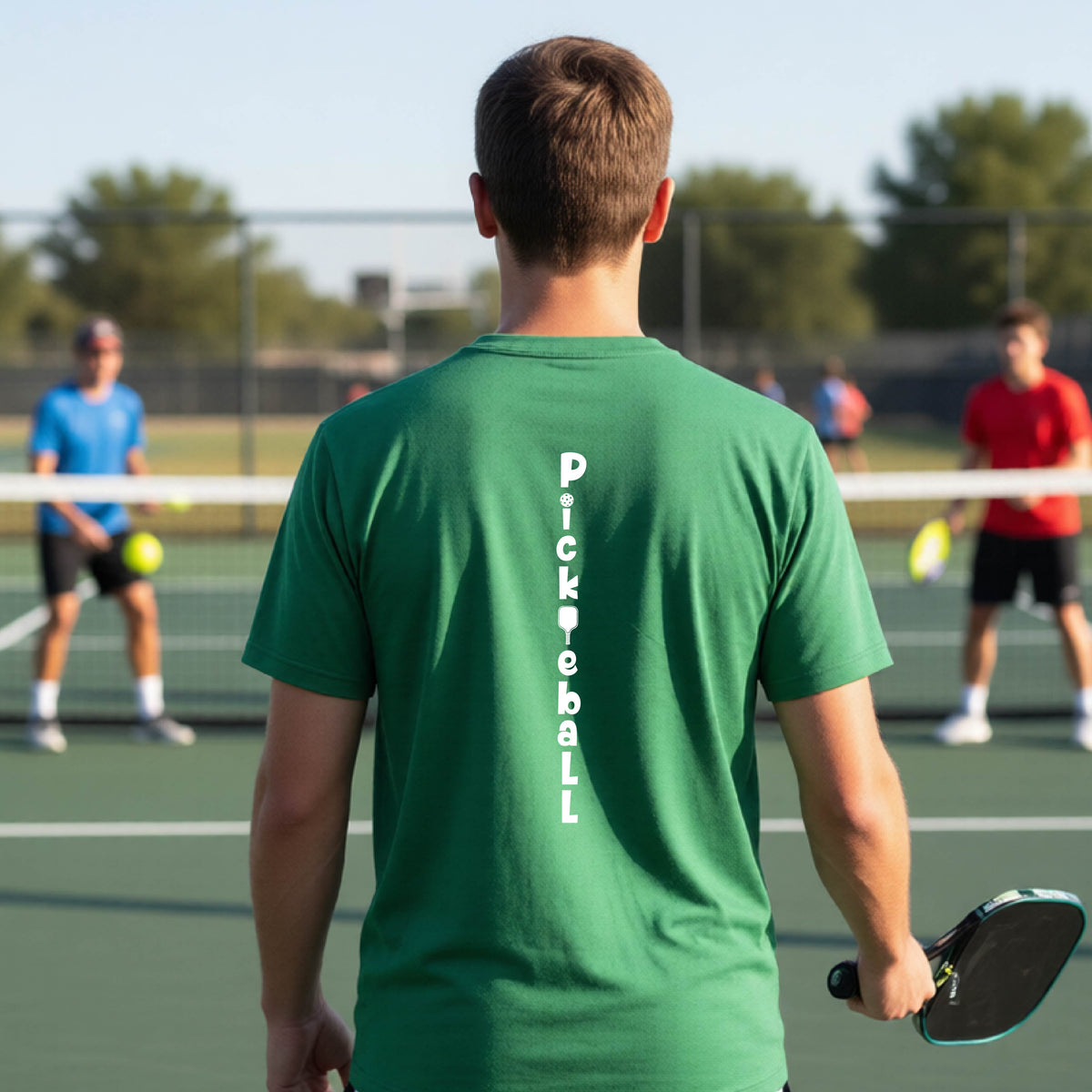 A young person wearing a green short-sleeve t-shirt with a white graphic text 'PICKLEBALL' on the back with a pickleball paddle replacing the L, standing on a pickleball court.