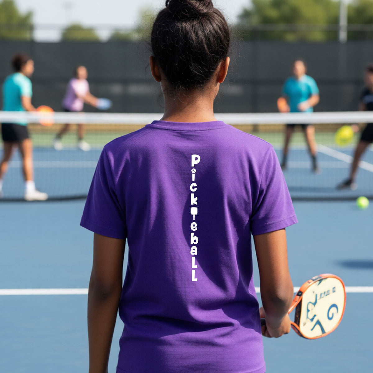 A young person wearing a purple short-sleeve t-shirt with a white graphic text 'PICKLEBALL' on the back with a pickleball paddle replacing the L, standing on a pickleball court.