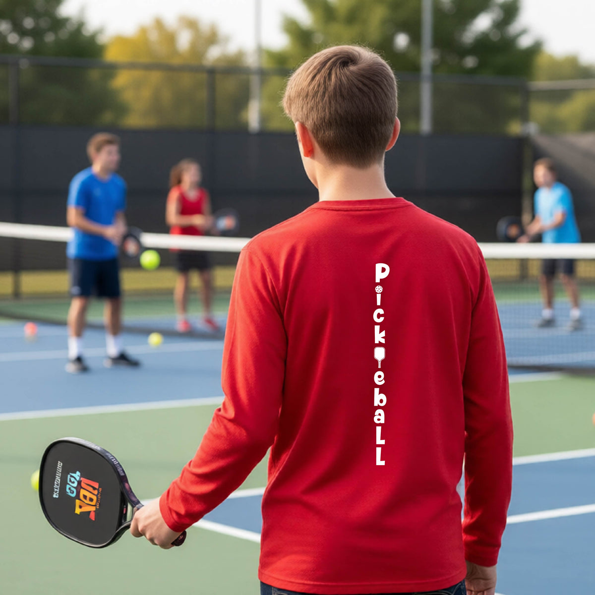 A young person wearing a red short-sleeve t-shirt with a white graphic text 'PICKLEBALL' on the back with a pickleball paddle replacing the L, standing on a pickleball court.