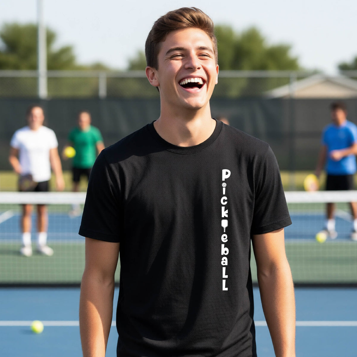 A young person wearing a black short-sleeve t-shirt with a white graphic text 'PICKLEBALL' on the front with a pickleball paddle replacing the L, standing on a pickleball court.