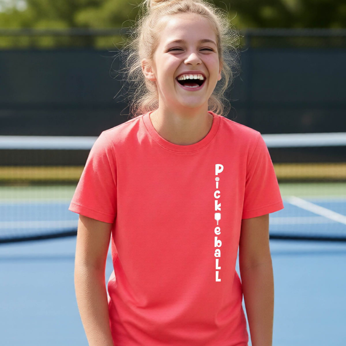 A young person wearing a coral-colored short-sleeve t-shirt with a white graphic text 'PICKLEBALL' on the front with a pickleball paddle replacing the L, standing on a pickleball court.