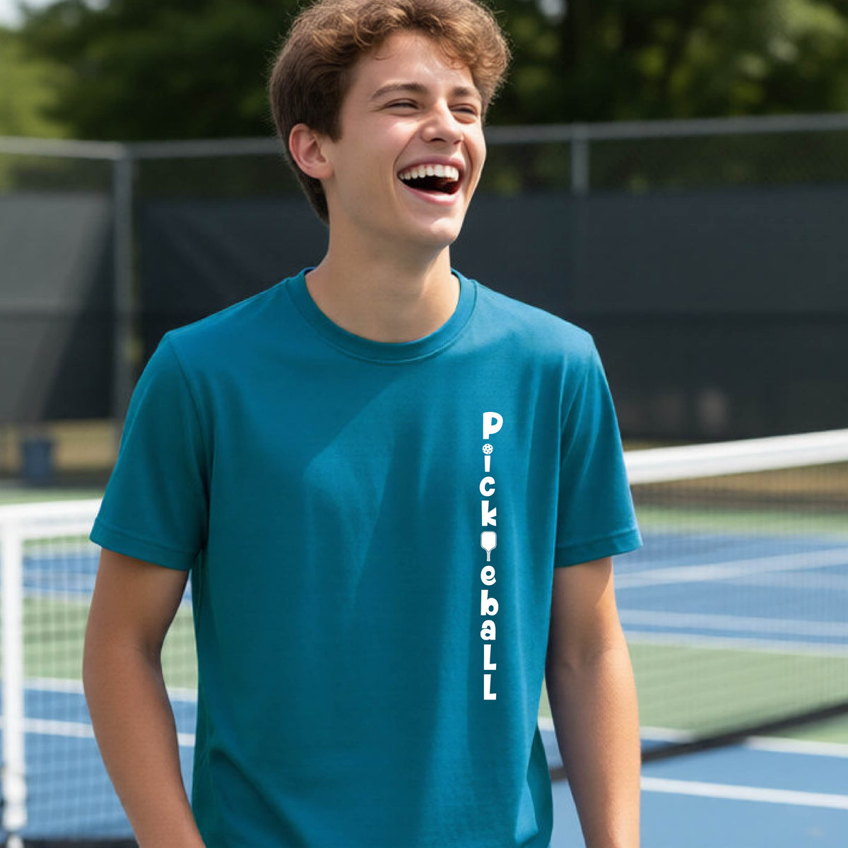 A young person wearing a teal short-sleeve t-shirt with a white graphic text 'PICKLEBALL' on the front with a pickleball paddle replacing the L, standing on a pickleball court.