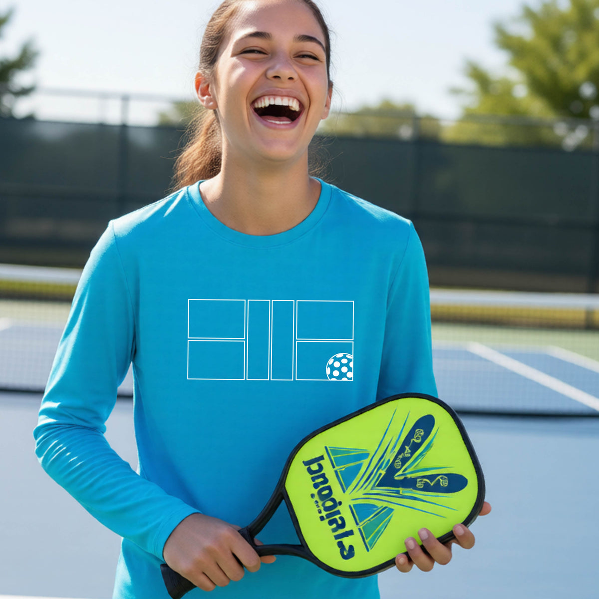 A young person laughing on a pickleball court, wearing a bright blue long-sleeve t-shirt with a white geometric graphic on the front of pickleball court and pickleball.