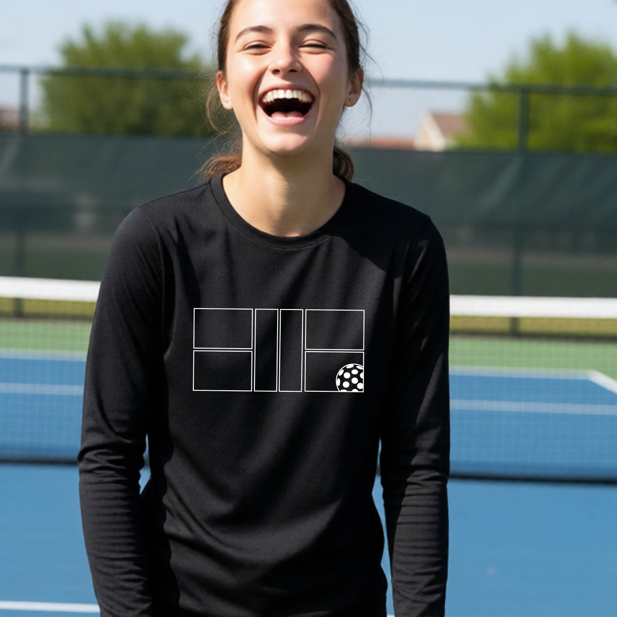 A young person laughing on a pickleball court, wearing a black long-sleeve t-shirt with a white geometric graphic on the front of pickleball court and pickleball.