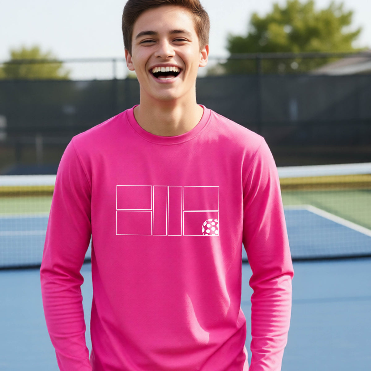 A young person laughing on a pickleball court, wearing a pink long-sleeve t-shirt with a white geometric graphic on the front of pickleball court and pickleball.