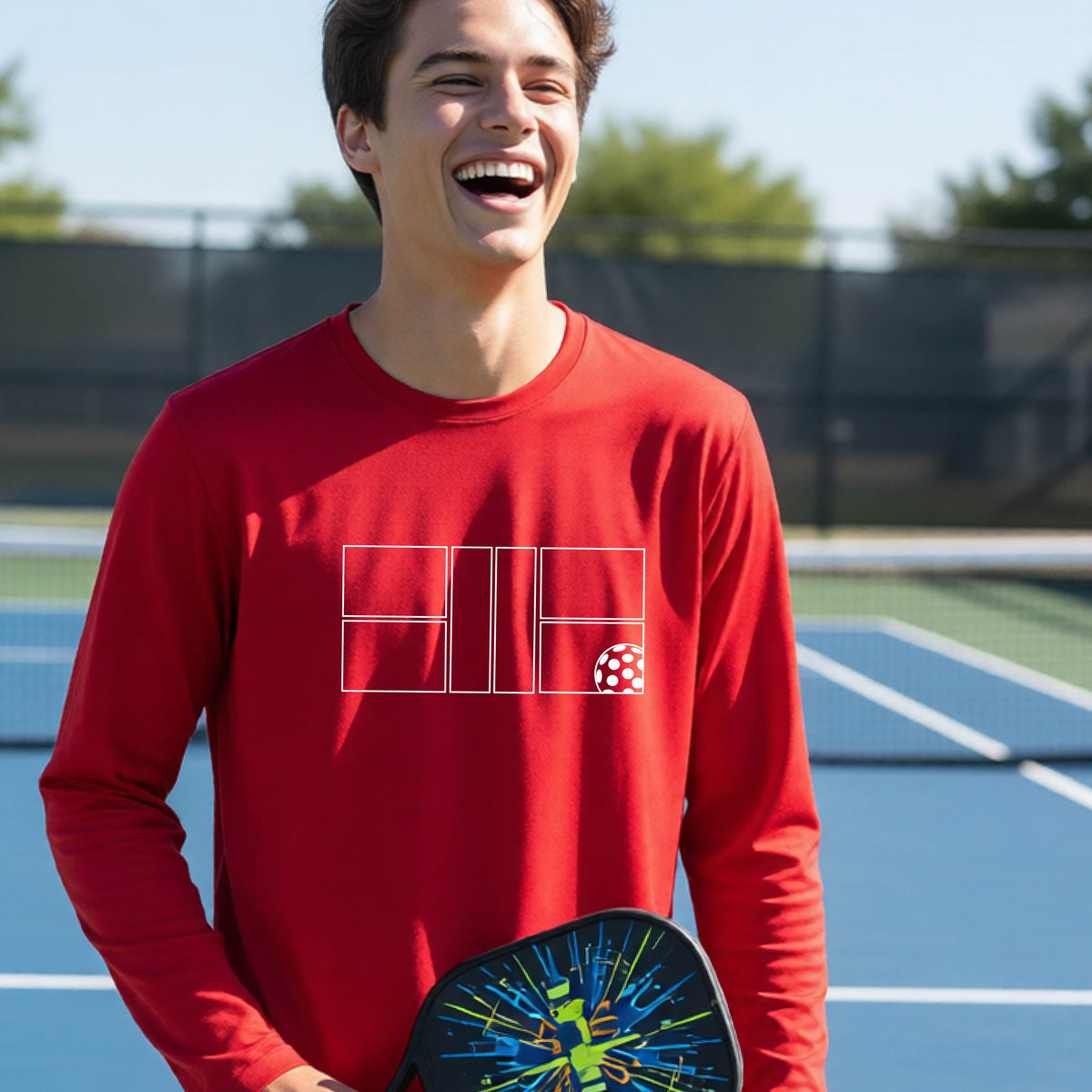 A young person laughing on a pickleball court, wearing a red long-sleeve t-shirt with a white geometric graphic on the front of pickleball court and pickleball.