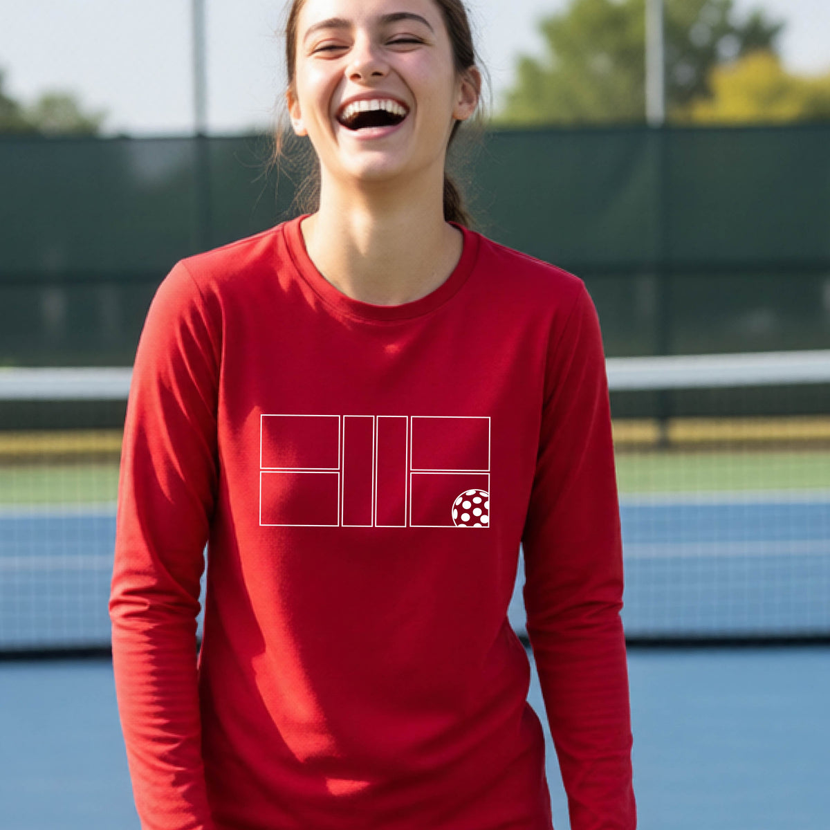 A young person laughing on a pickleball court, wearing a red long-sleeve t-shirt with a white geometric graphic on the front of pickleball court and pickleball.