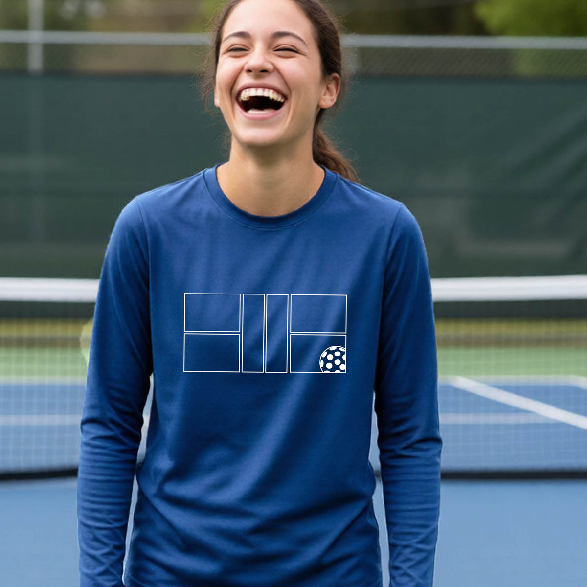 A young person laughing on a pickleball court, wearing a royal blue long-sleeve t-shirt with a white geometric graphic on the front of pickleball court and pickleball.