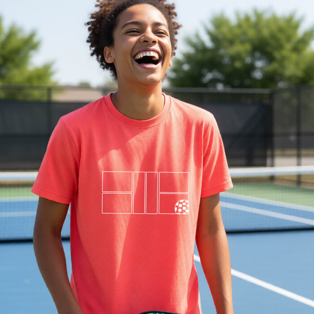 A young person laughing on a pickleball court, wearing a coral pink short-sleeve t-shirt with a white geometric graphic on the front of pickleball court and pickleball.