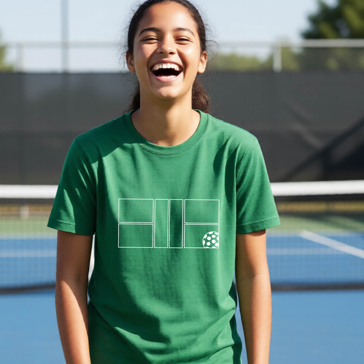 A young person laughing on a pickleball court, wearing a green short-sleeve t-shirt with a white geometric graphic on the front of pickleball court and pickleball.