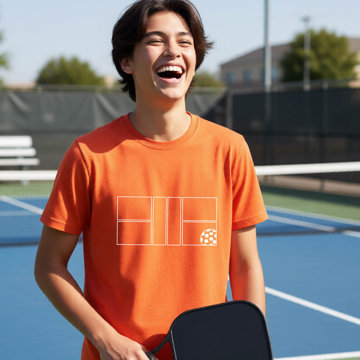 A young person laughing on a pickleball court, wearing a bright orange short-sleeve t-shirt with a white geometric graphic on the front of pickleball court and pickleball.