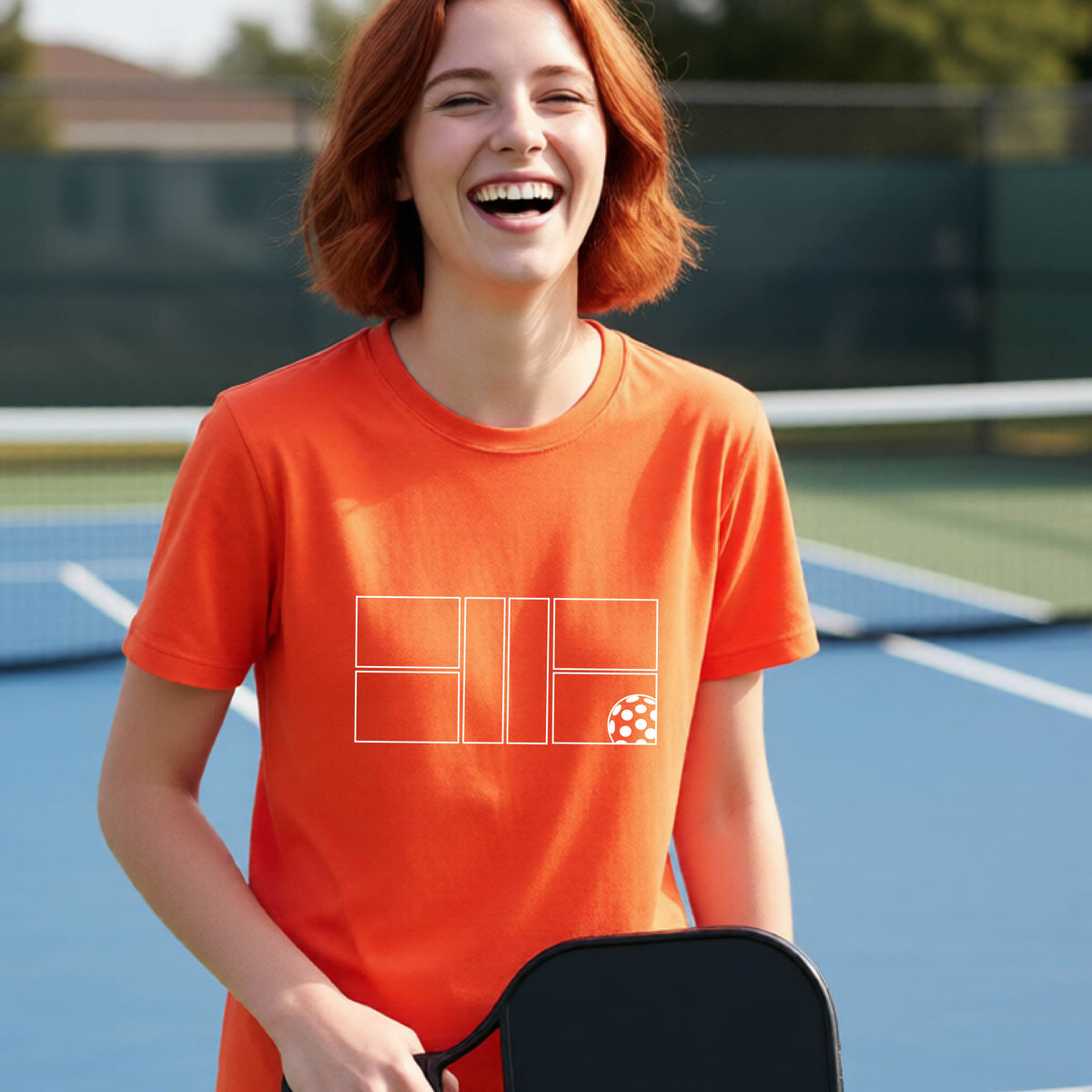 A young person laughing on a pickleball court, wearing a bright orange short-sleeve t-shirt with a white geometric graphic on the front of pickleball court and pickleball.