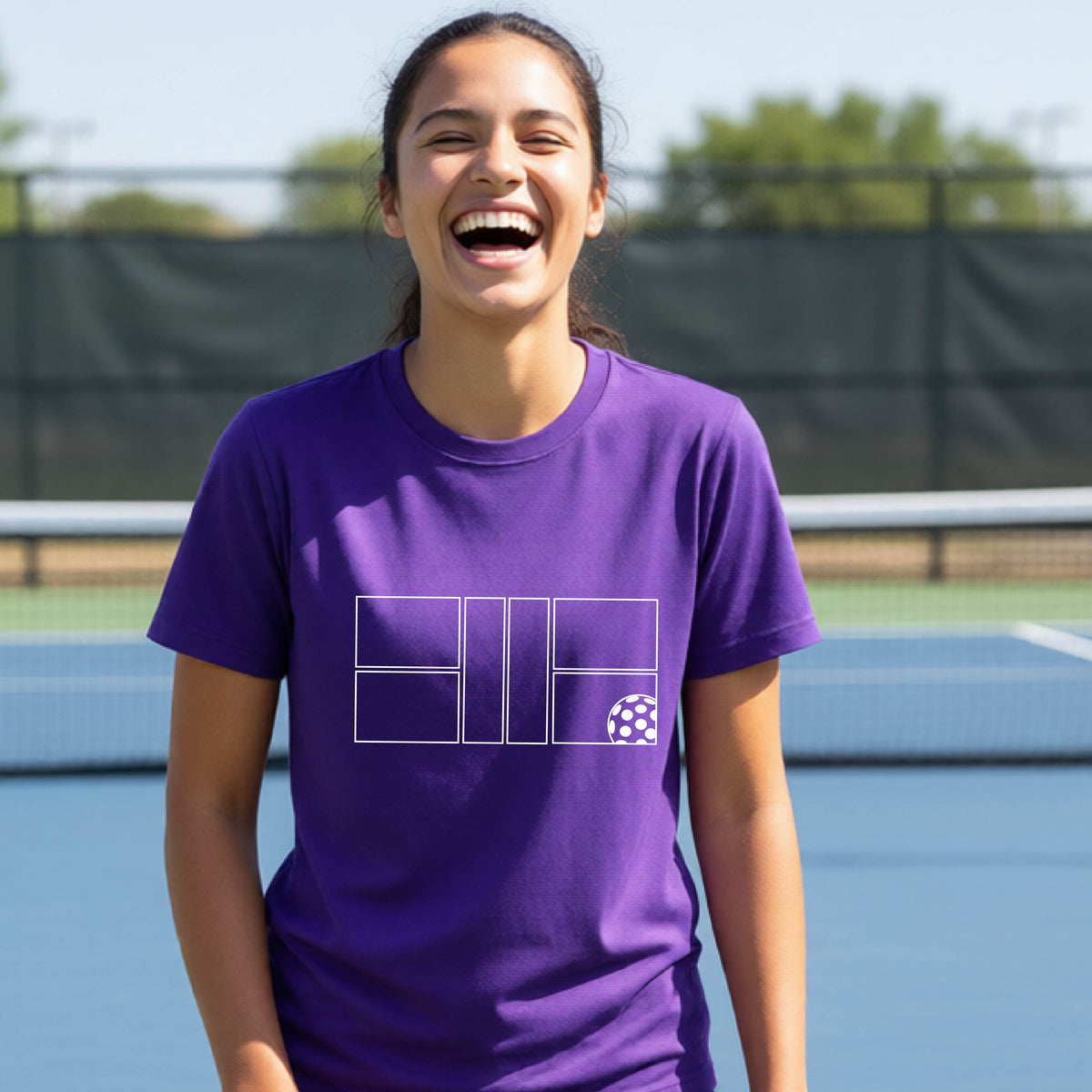 A young person laughing on a pickleball court, wearing a purple short-sleeve t-shirt with a white geometric graphic on the front of pickleball court and pickleball.