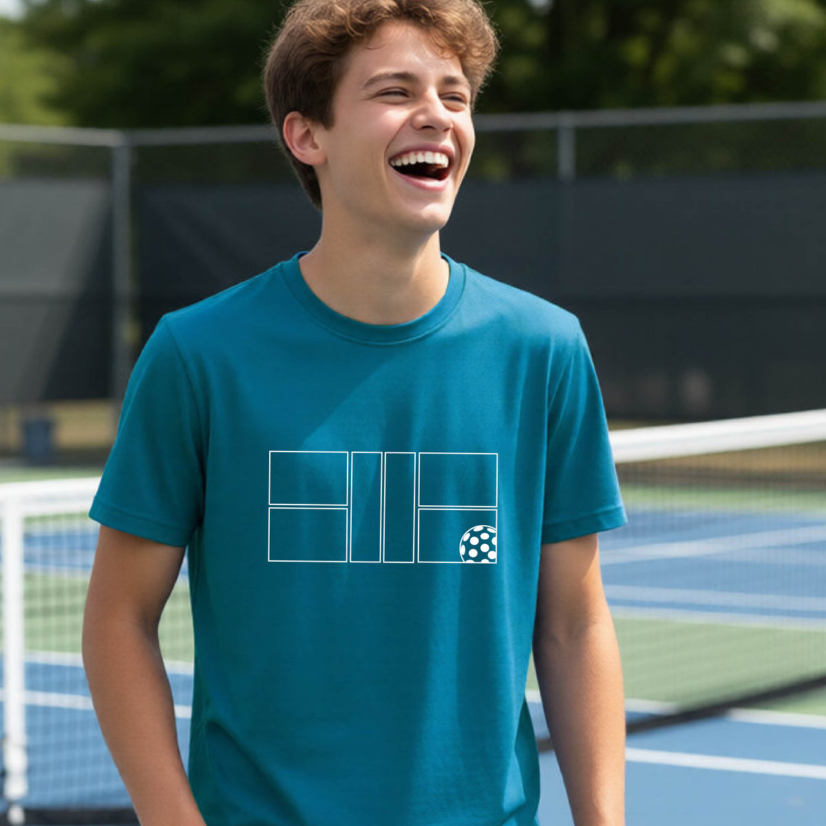 A young person laughing on a pickleball court, wearing a teal short-sleeve t-shirt with a white geometric graphic on the front of pickleball court and pickleball.