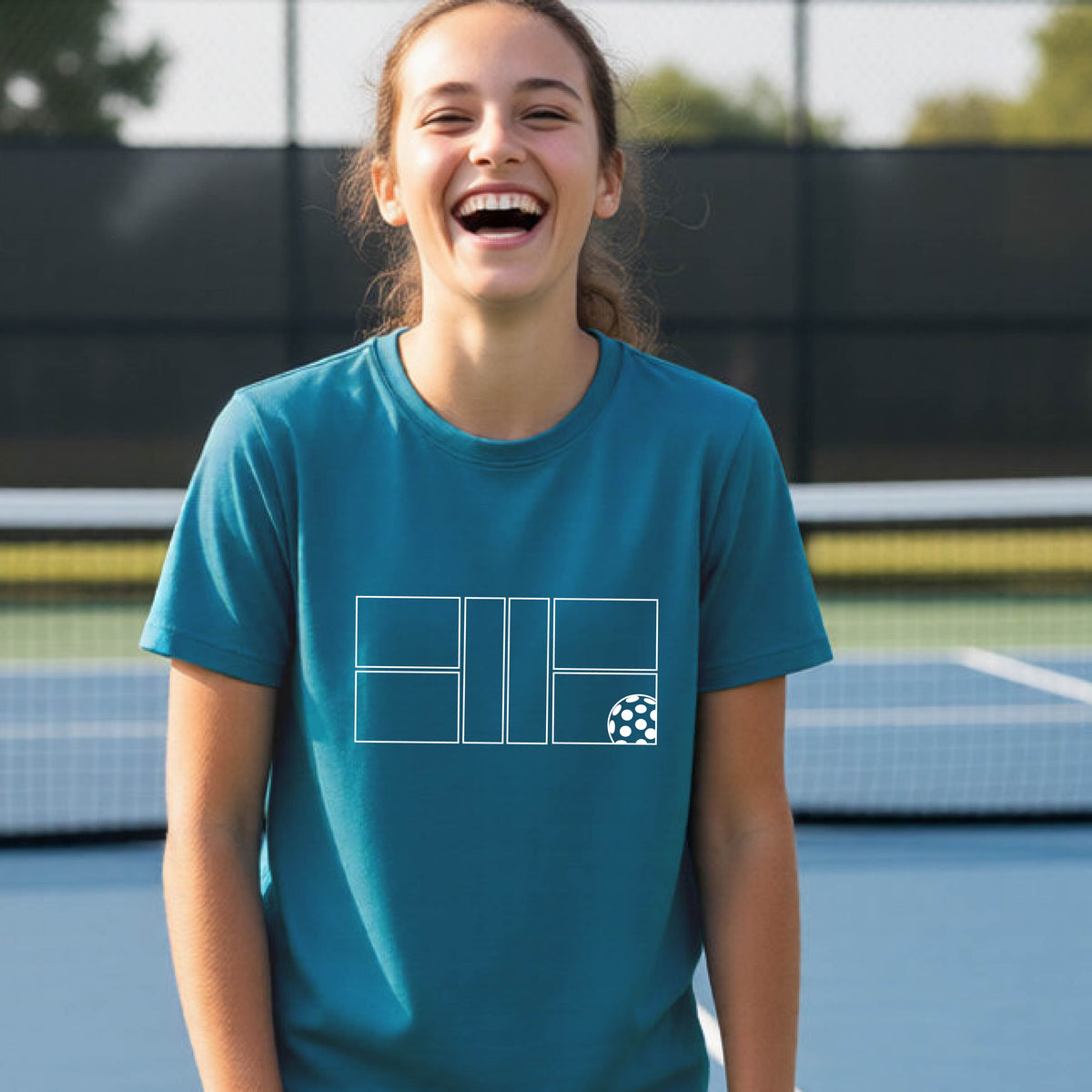 A young person laughing on a pickleball court, wearing a teal short-sleeve t-shirt with a white geometric graphic on the front of pickleball court and pickleball.