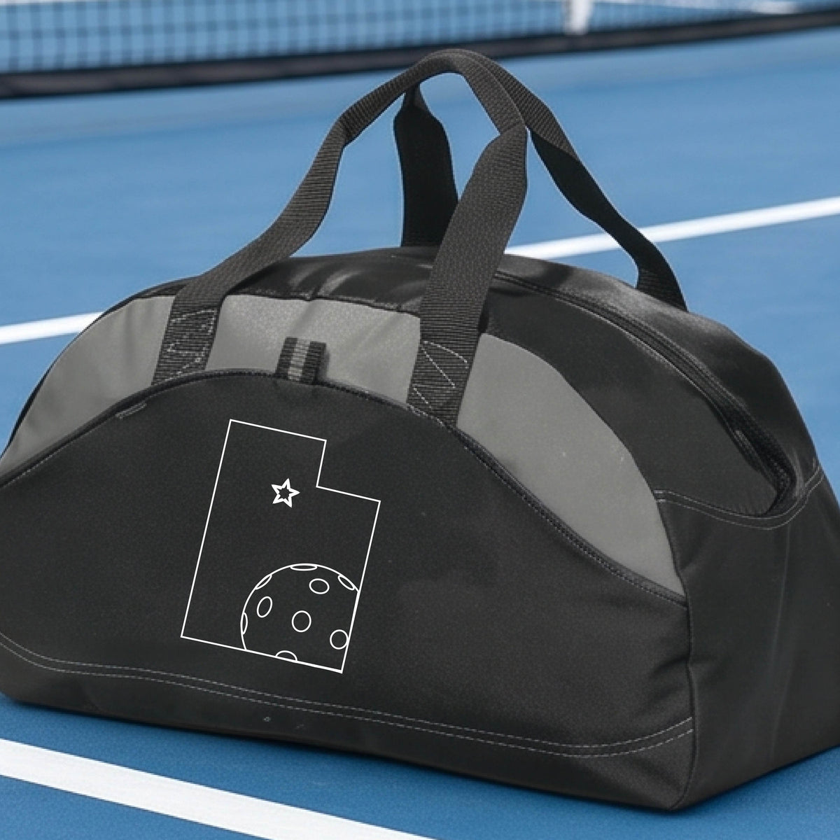 A close-up of a medium-size black with gray panels duffel bag, with a white graphic of the State of Utah , including an outline of a white pickleball in the lower right corner and a star outline in the center of the graphic, on the front of the bag