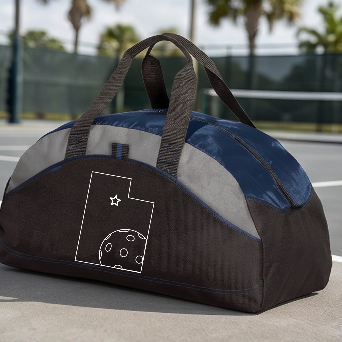 A close-up of a medium-size navy blue, black with gray panels duffel bag, with a white graphic of the State of Utah , including an outline of a white pickleball in the lower right corner and a star outline in the center of the graphic, on the front of the bag