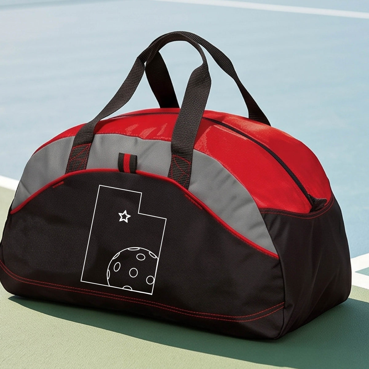 A close-up of a medium-size red, black with gray panels duffel bag, with a white graphic of the State of Utah , including an outline of a white pickleball in the lower right corner and a star outline in the center of the graphic, on the front of the bag