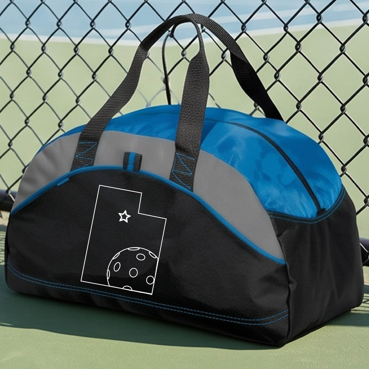 A close-up of a medium-size black and royal blue with gray panels duffel bag, with a white graphic of the State of Utah , including an outline of a white pickleball in the lower right corner and a star outline in the center of the graphic, on the front of the bag