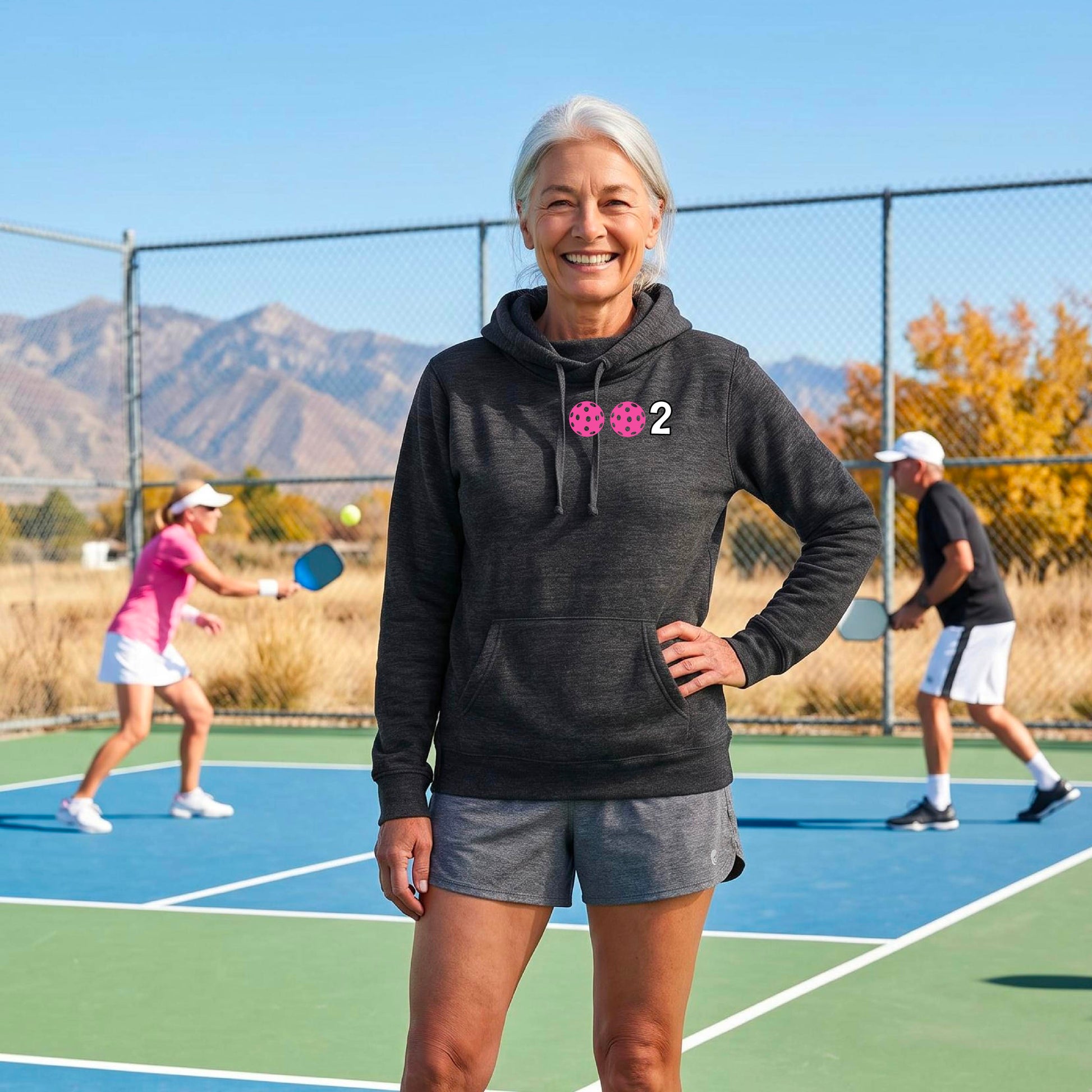 A woman smiling outdoors on a pickleball court, wearing a heather black hoodie with two pickleballs and the number 2 printed on the chest.